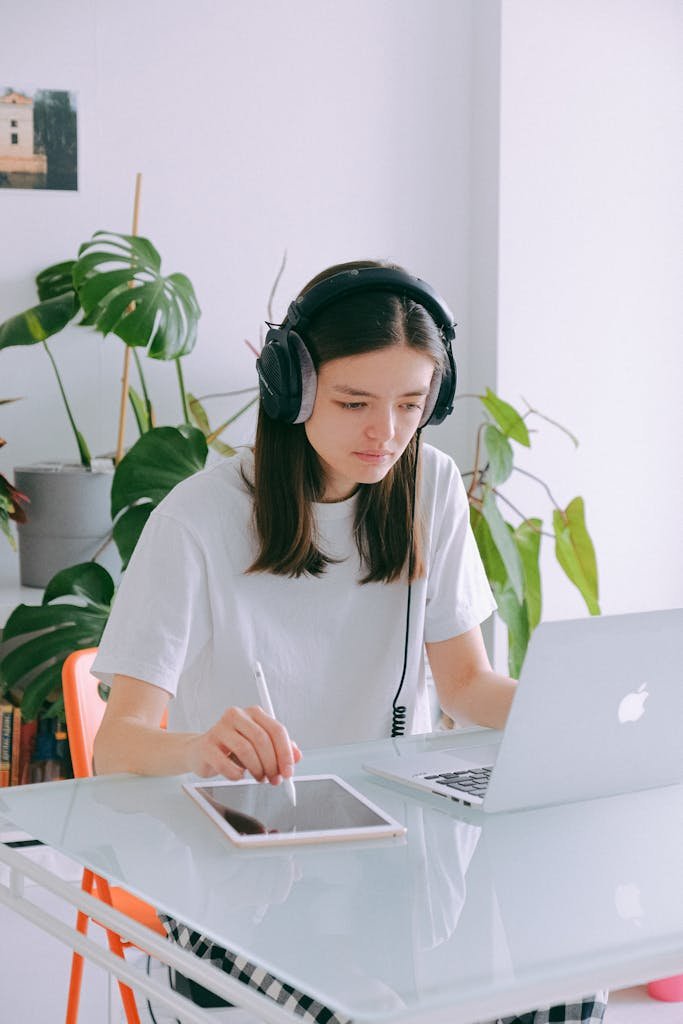 Young woman working from home, using laptop, tablet, and headphones at a minimalist desk.